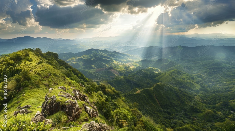 Obraz premium A vibrant green mountain ridge dominates the foreground as powerful sun rays descend from cloudy skies onto the distant, hazy valleys below.