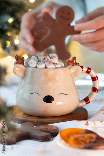 A mug of hot chocolate top with marshmallow and cacao powder with candied orange, cozy Christmas decoration while hands holding chocolate man with light bokeh in background.