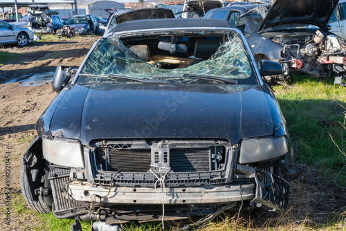 Photos a black convertible with a completely shattered windshield and missing front bumper, parked on a grassy patch in a mud-strewn salvage yard