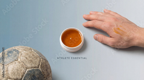 An athlete's hand applies a golden balm for muscle recovery after a football game. The balm is in a jar and a ball is beside.