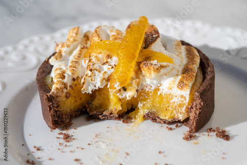 a lemon meringue tartlet with a dark chocolate crust, featuring a bite taken out to reveal the gooey yellow filling, topped with toasted meringue and candied peel on a white plate.