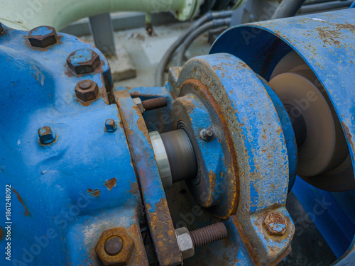 Close-up of a grease fitting (zerk) on rotating industrial machinery. Visible rust, bolts, and motion blur highlight active lubrication in a heavy-duty environment.
