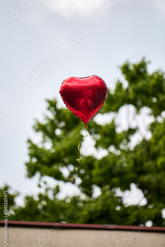 Red heart-shaped balloon floating above green tree canopy outdoors