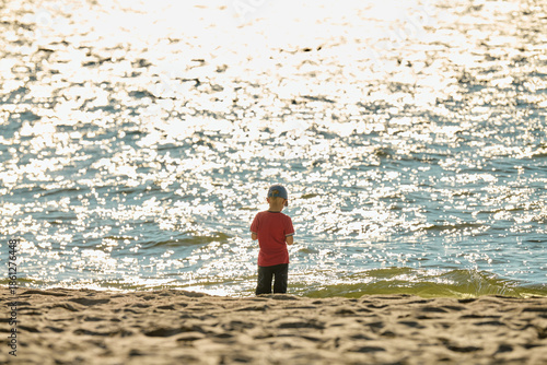 Young child standing on sunlit sandy beach facing sparkling water
