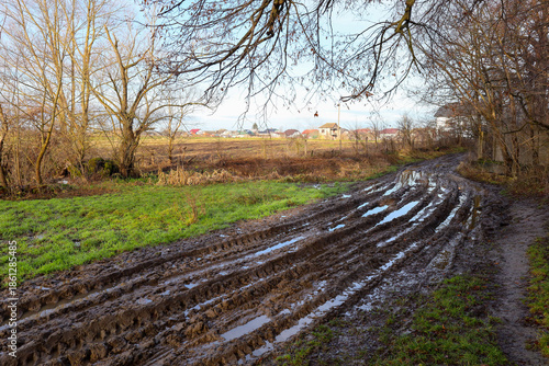 A muddy dirt road with deep ruts and puddles passes through a picturesque countryside in autumn or spring. The panorama includes cozy village houses on the horizon, bare tree branches in the foregroun