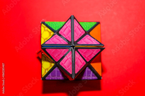 A complex multifaceted geometric puzzle with vibrant textured panels of pink, green, and purple colors set against a rich red background. This intellectual toy stimulates logical thinking,