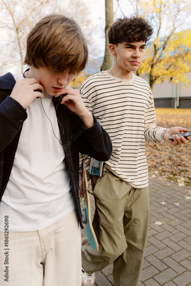 Obraz premium Two teenagers stroll through a park filled with autumn leaves, one adjusting his headphones while the other casually checks his phone. The trees around them are beautifully colored.