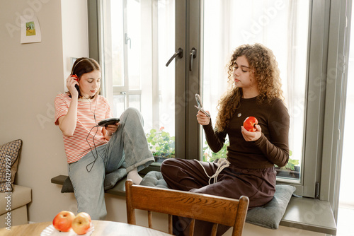 Fotografija Two friends relax together by a sunny window, listening to music and enjoying fresh apples