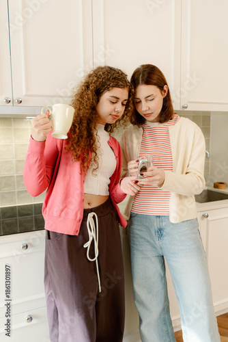 Two cheerful friends share a lighthearted moment in a bright kitchen, one holding a cup and the other examining a camera. Their smiles show a bond forged over shared passions.