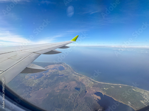 Commercial aircraft wing seen through window, high-altitude view of ocean and shore, bright daylight, aviation, tourism and travel concept.