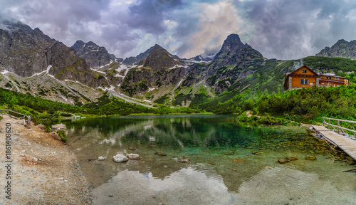 A dramatic landscape shot of a hiking path at the edge of Green Lake (Zelene pleso). Massive granite peaks with snow patches are shrouded in heavy grey storm clouds. Tatra National Park, Slovakia.