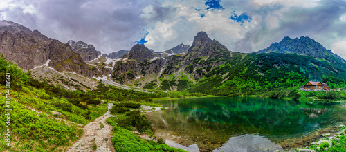 A dramatic landscape shot of a hiking path at the edge of Green Lake (Zelene pleso). Massive granite peaks with snow patches are shrouded in heavy grey storm clouds. Tatra National Park, Slovakia.