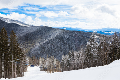 Snowy mountains stretch beneath a partly cloudy sky, with ski lift tracing slope beside frosted evergreens. Serene winter scene evokes alpine solitude and outdoor adventure