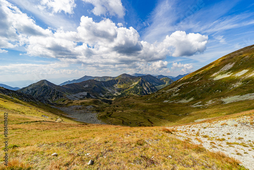 A narrow rocky path winds up a steep grassy slope toward a mountain summit. Scenic view from Hladke sedlo in High Tatras National Park, featuring a turquoise lake in the valley under a cloudy blue sky