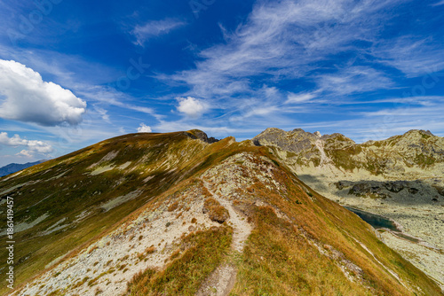A narrow rocky path winds up a steep grassy slope toward a mountain summit. Scenic view from Hladke sedlo in High Tatras National Park, featuring a turquoise lake in the valley under a cloudy blue sky