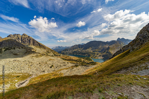 A narrow rocky path winds up a steep grassy slope toward a mountain summit. Scenic view from Hladke sedlo in High Tatras National Park, featuring a turquoise lake in the valley under a cloudy blue sky