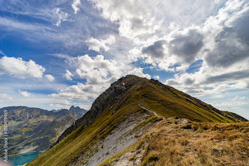 A narrow rocky path winds up a steep grassy slope toward a mountain summit. Scenic view from Hladke sedlo in High Tatras National Park, featuring a turquoise lake in the valley under a cloudy blue sky
