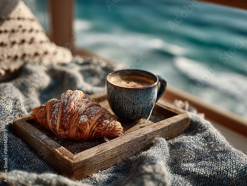 a cozy setting with a wooden tray holding a croissant and a cup of coffee on a knitted blanket. In the background, there is a view of the ocean, adding a serene and relaxing atmosphere to the scene.