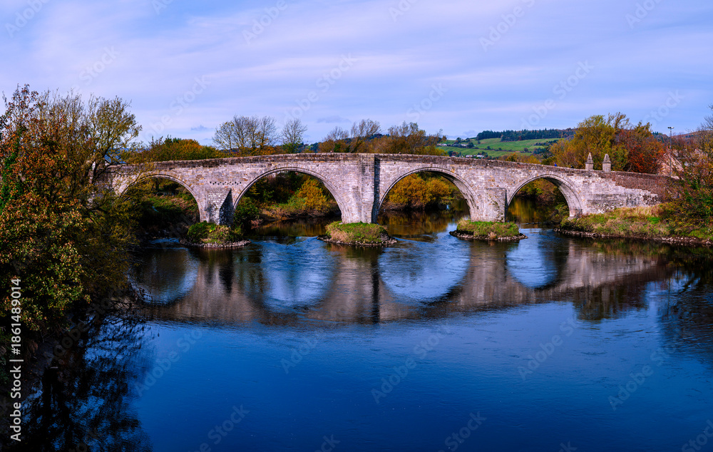 Fototapeta premium Stirling Old Bridge spanning the River Forth with the National Wallace Monument rising beyond, a tranquil and vibrant autumn landscape and the landmark historic architectures of Scotland in UK