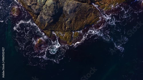 Rough waves dance around weathered stones, glistening under a clear sky. The scene captures nature's beauty at the shore, filled with life and movement.