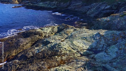Rugged rocks jut out into the crystal-clear blue water, where gentle waves lap against the stony shore under a bright sky. Nature's beauty shines in every detail.
