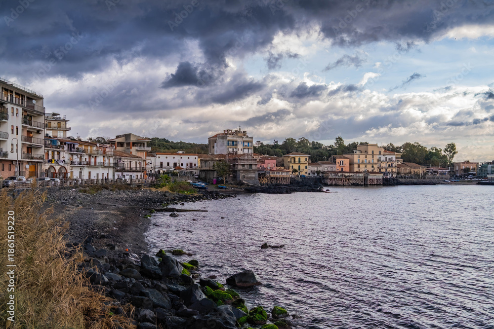 Fototapeta premium A wide-angle shot of the picturesque coastal village of Capomulini, Acireale, in Sicily, Italy, captured on an overcast day. The image features traditional Mediterranean buildings lining the rocky, vo