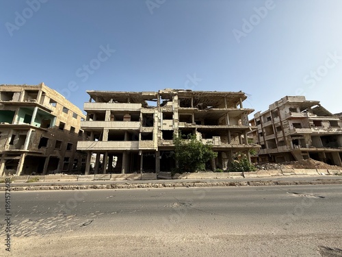 Photos War-torn buildings, ruined architecture, and damaged cityscape under a clear sky, suggesting conflict or disaster zone