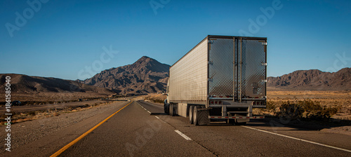 Following behind a white truck on a two lane highway in the desert with a rocky hill in the distance. The image is from the I-15 in Nevada