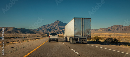Following behind a white truck and trailer and a white pick up truck  on a two lane highway in the desert . The image is from the I-15 in Nevada