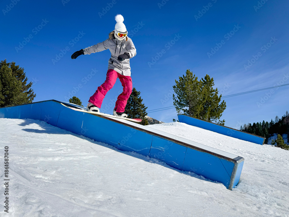 Fototapeta premium Freestyle snowboarder in pink pants slides a blue box rail in a terrain park. Young woman is doing a stylish boardslide trick during a fun lap through snowpark at Austrian ski resort on a sunny day.