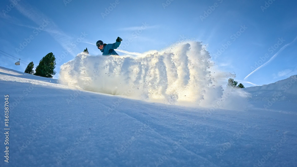 Obraz premium LENS FLARE, LOW ANGLE VIEW, SILHOUETTE: Snowboarder sends a cloud of snow towards camera as he makes a turn while riding down the ski slope. Fun snowboarding at ski resort in the snowy Austrian Alps.