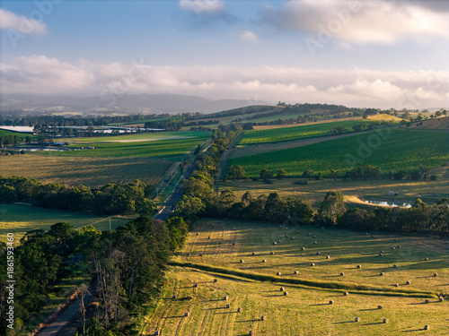 Принти на полотні Golden hour illuminates a vast agricultural landscape featuring hay bales, green fields, a winding road, and distant misty hills