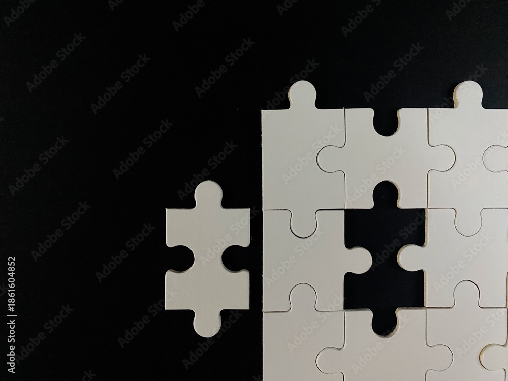 Fototapeta premium Minimalist top-down view of a white jigsaw puzzle on a black background. One piece is separated, symbolizing the final step, solution, and business completion.