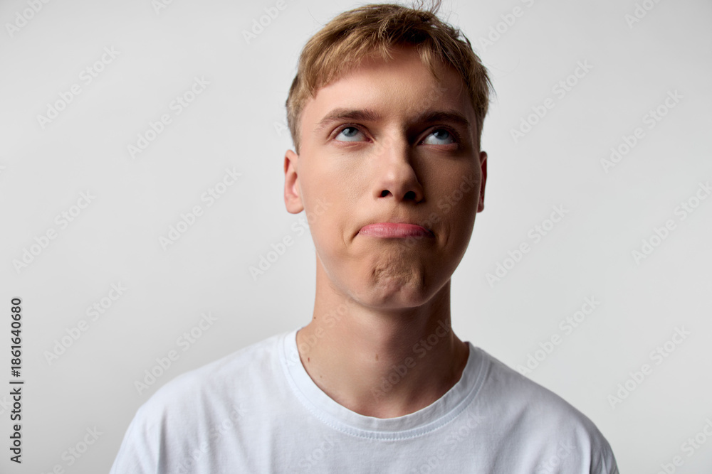 Obraz premium Young man expression thinking pensive confusion gaze upward look white t shirt studio lighting calm mood emotion image candid minimal background neutral tones indoor light focus on facial features