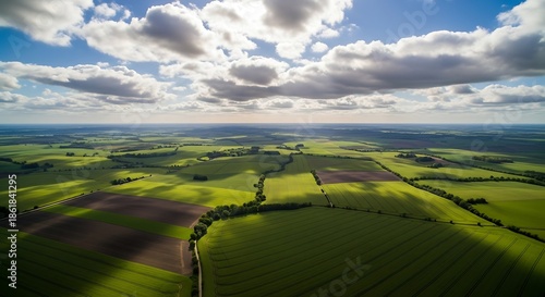 An aerial shot captures sunlit fields and shadows under a partly cloudy sky. The landscape stretches towards the horizon