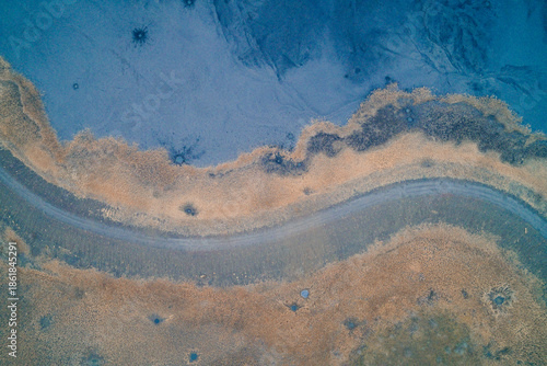 Curved dirt road separating toxic blue sludge and dry grass, surreal industrial pollution texture and landscape background, aerial top view drone shot