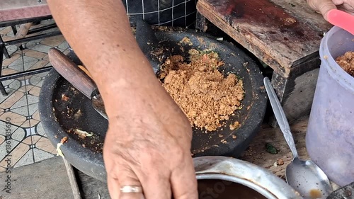 A vendor's hands grinding fresh ingredients in a stone mortar (cobek) to prepare traditional Indonesian Lotek peanut sauce. Authentic street food preparation video.