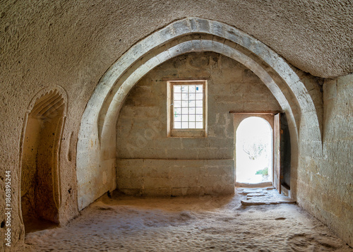 Interior of a rock-cut mosque in Zelve Valley Open Air Museum, Cappadocia, Turkey. Features ancient stone architecture with arched ceilings, a gridded window, and an open doorway.