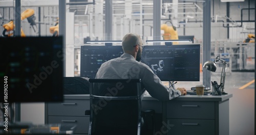 Engineers Monitoring Automation Model. Man Works Intensely at Double Monitor, Where 3D Model Robot and Data Metrics Displayed. Control Room Overlooks Modern Factory Floor. Robotics Control Room.