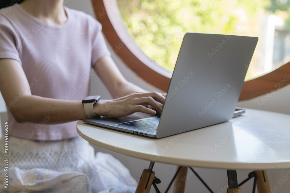 Fototapeta premium A person typing on a laptop at a modern table near a circular window.