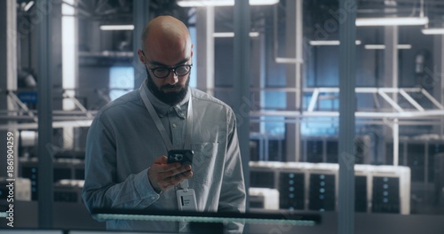 Bearded Male IT Engineer in Glasses Stands and Uses Smartphone to Check Critical Notifications or System Status Large Scale of Data Center. Emphasizes Mobile Management and Operational Urgency.