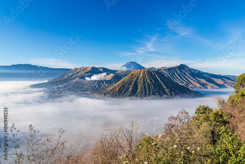 Morning view, Mount Bromo, East Java, Indonesia. Smoke rising from one of the active volcanoes, Fog covering the surrounding desert. Blue sky above. 
