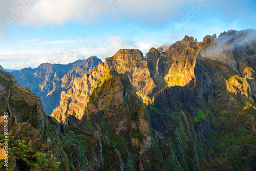 Stunning Pico do Arieiro mountain landscape in Madeira, Portugal, jagged peaks and hiking trail above clouds. Morning sun dramatically highlights rugged rock faces and green slopes of island massif