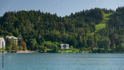 Bled, Slovenia - August 11, 2025: A panoramic view showcases the lakeside hotels and private villas of Bled, Slovenia, nestled against a steep, densely forested alpine hillside under a clear sky.