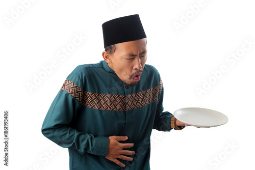 Asian Muslim man wearing a teal koko shirt (baju koko) and black peci hat, looking nauseous while holding a white empty plate after overeating during Ramadan fasting, isolated on a white background.