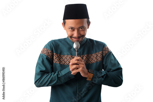 Indonesian Muslim man wearing a green koko shirt and a black peci cap (songkok), holding a spoon with both hands while looking at the camera with hungry expression, Isolated on a white background