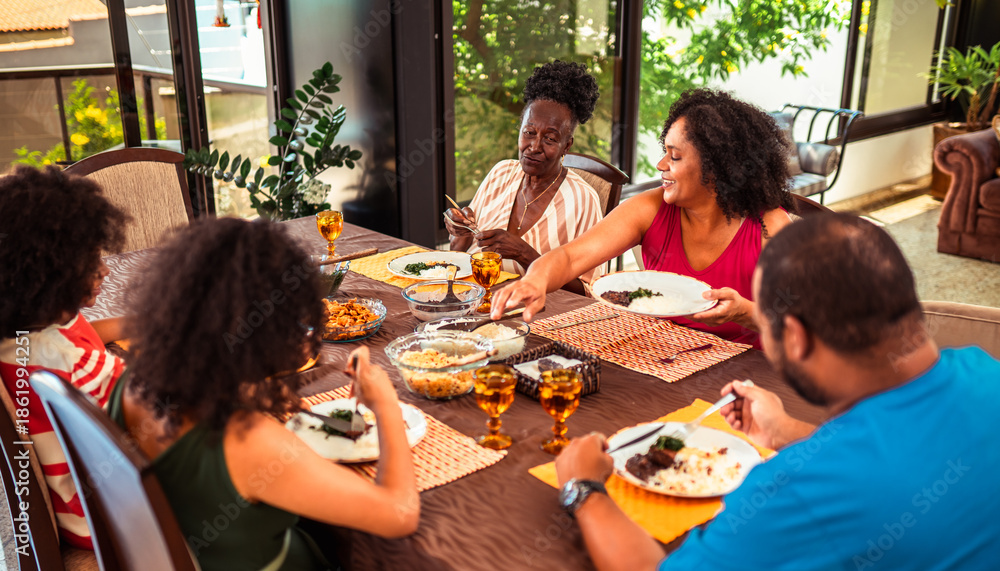Obraz premium Happy multi-generational Black family enjoying a homemade meal together at a dining table, sharing food and laughter in a bright home setting.
