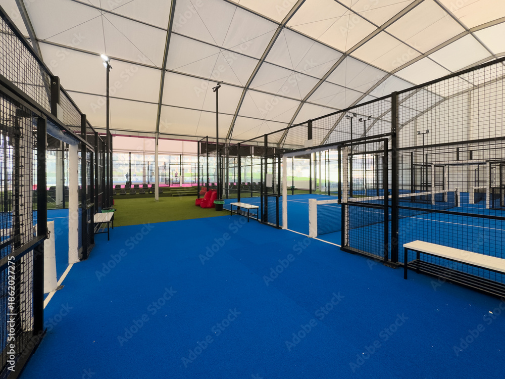 Obraz premium Spacious indoor padel court featuring a vivid blue surface, surrounded by metal fences and overhead bright lights, perfect for training or competitive matches within a well-maintained sports facility.