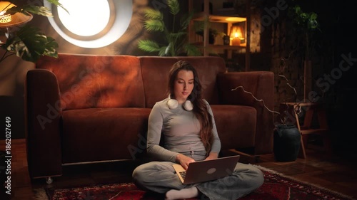 Young woman sitting on the floor at home using a laptop. Cozy living room, warm light, casual lifestyle, remote work, online study, freelance work, calm evening atmosphere