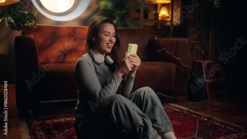 Happy young woman sitting on the floor at home reacts with joy to exciting news on her smartphone. Cozy living room, warm light, emotional moment, lifestyle, positive emotions, communication